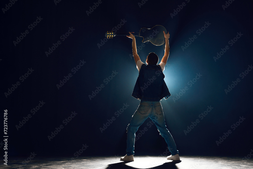 Rear view portrait of man in denim outfit holds electric guitar under head stands on stage. Silhouette of Rock vocalist. Concept of Rock-n-roll, music and dance, festivals and concerts, culture. Rear view portrait of man in denim outfit holds electric guitar under head stands on stage. Silhouette of Rock vocalist. Concept of Rock-n-roll, music and dance, festivals and concerts, culture.