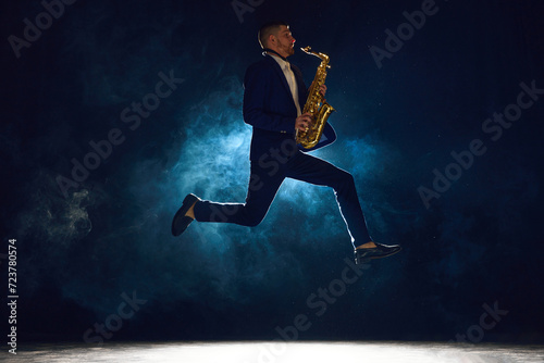 Dynamic shot of artistic man, solo performer jumping while playing jazz melody on saxophone against dark background with smoke. Concept of art, instrumental music, dance, culture, festivals, concerts
