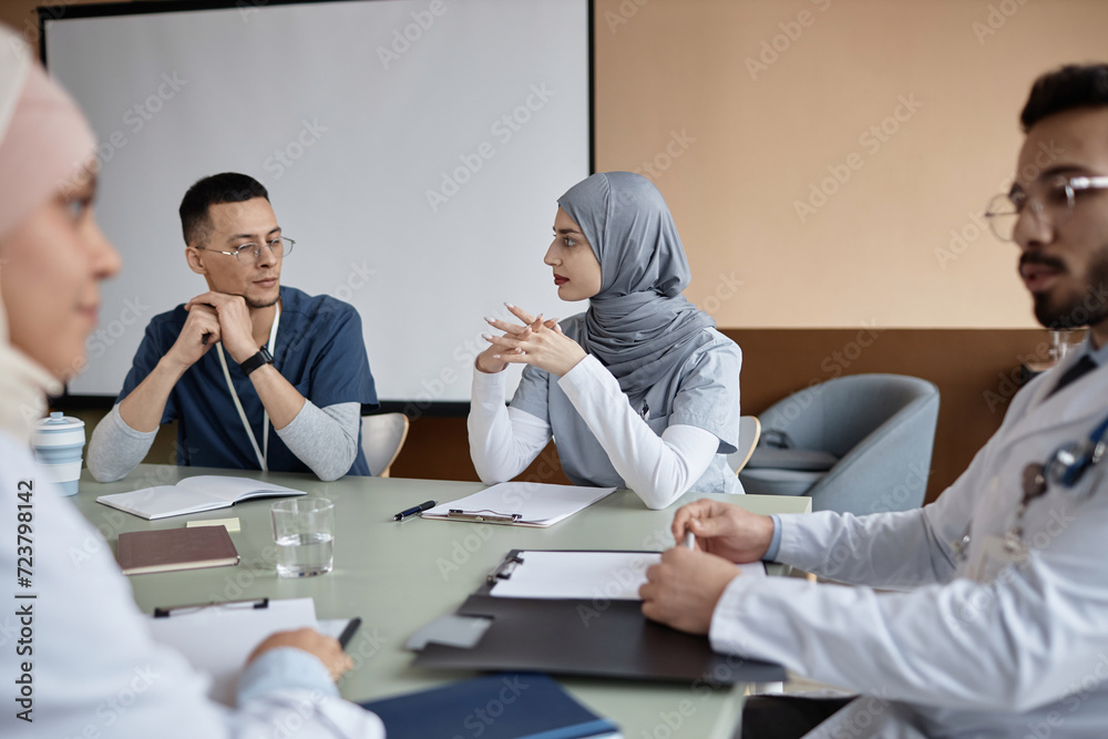 Fototapeta premium Islamic woman doctor wearing gray hijab speaking to male coworker sitting at table during conference