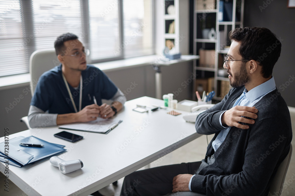 Side view of Middle Eastern man patient in glasses complaining of shoulder pains during visit with defocused male doctor in clinic Side view of Middle Eastern man patient in glasses complaining of shoulder pains during visit with defocused male doctor in clinic