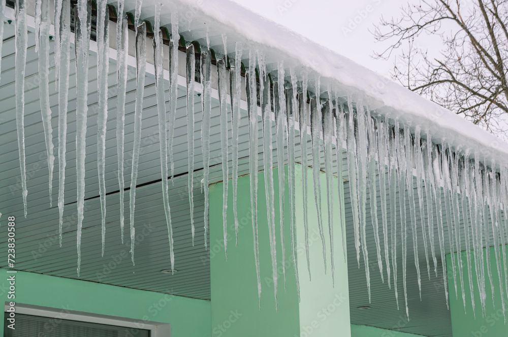 Icicles hang from the roof when the snow melts on the roof of the building. Long icicles grow with warming in spring. The icicles are dripping with water. Icicles are dangerous when falling on people.