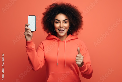 Young fitness woman, wearing a red tracksuit, holding a smartphone and showing thumb up gesture, isolated on red background. photo