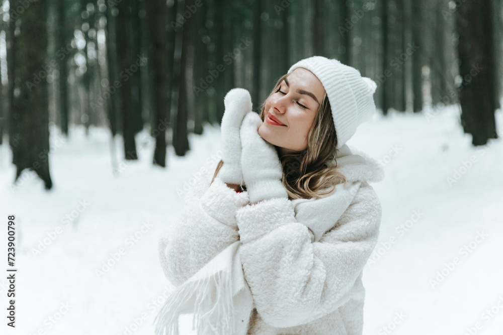 Young woman Enjoying snowy winter forest and warm clothes. Dressed white coat scarf hat and mittens fold palms to face