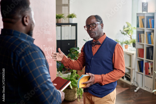 Portrait of Black college professor talking to student standing by blackboard in classroom