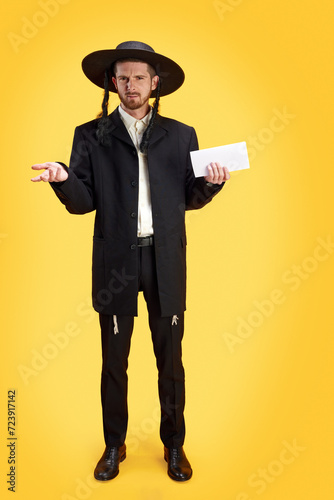 Skeptical Jewish man in traditional garb holding an envelope, evoking curiosity and intrigue against sunny yellow background. Purim, festival, holiday, celebration Pesach or Passover concept. Ad photo