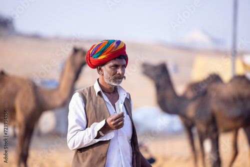 Portrait of an old male from rajasthan in traditional white dress and colourful turban with camel at desert fair ground during pushkar fair. photo