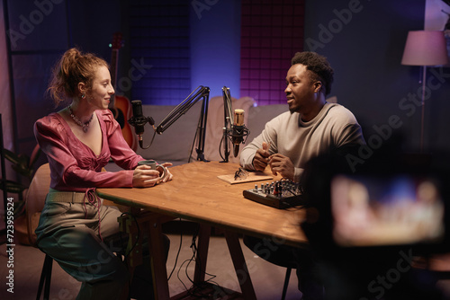 Medium long shot of trendy Caucasian girl and young African American man recording vlog sitting at table in podcast room with neon light