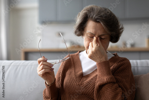 Exhausted elderly woman taking off glasses, suffering from blurry vision or dizziness, have eyesight problems, struggle with headache or migraine from high blood pressure photo