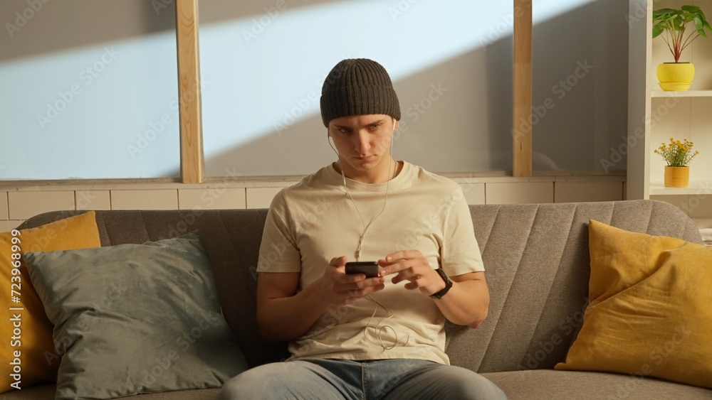Portrait of young person in the room sitting on the couch. Man in headphones listening to music on smartphone.