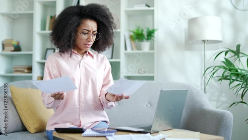 Confused young african american female having difficulty with paper work doing household budget sitting on sofa in living room at home office. Puzzled shocked black woman looks at large utility bills photo