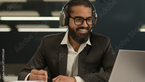 Happy smiling satisfied Arabian man business manager entrepreneur employer Indian positive smile businessman writing notes in notebook wear headphones online lesson class video call studying in office