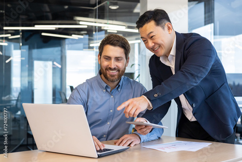 Two professional businessmen working together on a project in the office, discussing over a laptop and tablet with enthusiasm.