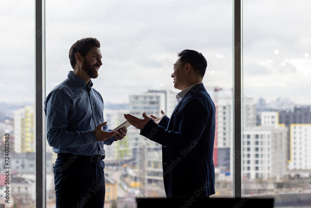 Two businessmen in silhouette converse in a high-rise office, reflecting teamwork, strategy, and corporate partnership. Two businessmen in silhouette converse in a high-rise office, reflecting teamwork, strategy, and corporate partnership.