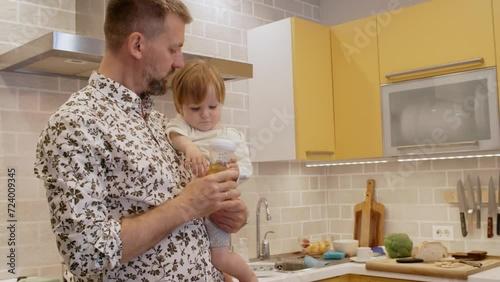 Tilt up of caring bearded father in stylish shirt standing in kitchen and holding toddler girl, giving her baby bottle with diluted juice photo