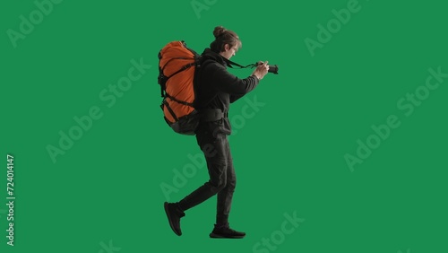 A male traveler takes pictures with a camera while hiking. A tourist with a backpack on his back stands full length in a studio on a green screen. Concept of travel, active rest, hiking. Side view.