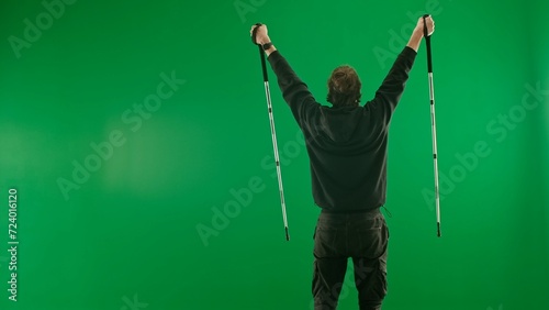 A young tourist joyfully makes a victory gesture, raising his hands in the air. A man with trekking poles in his hands stands in the studio on a green screen. Back view. © kinomaster