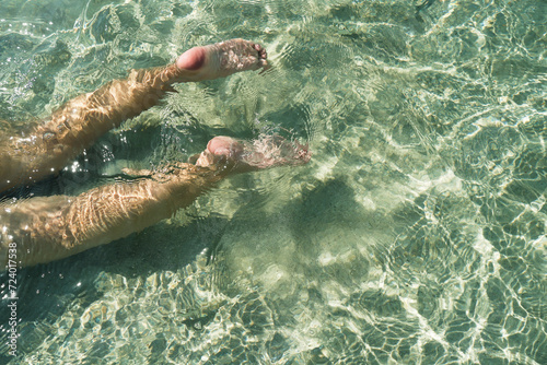Woman swimming on the sea. Female legs under clear sea water. Realistic top view photo of a women's legs barefoot. Relax woman lying in turquoise crystalline blue water on beach vacation summer.