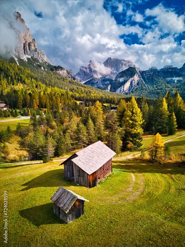 Aerial drone view of Dolomites Mountains house and forest in autumn season. Blue sky with clouds. Italy