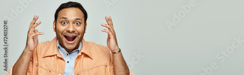 jolly surprised indian man in orange jacket smiling straight at camera on gray background, banner photo