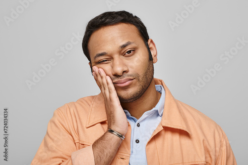 tired handsome indian man in orange vivid jacket posing with hand on face on gray background