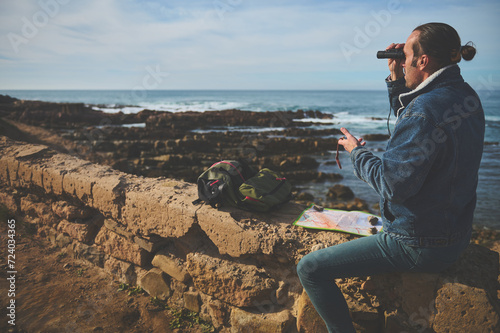 Young adventurer holding compass and using binoculars, looking through to the distance, sitting on the rock by Ocean