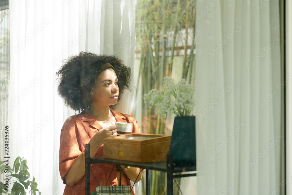 Pensive Latin woman drinking cup of coffee during break and looking outside through window Pensive Latin woman drinking cup of coffee during break and looking outside through window
