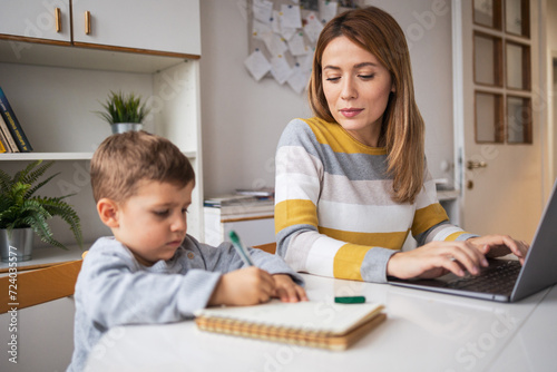 Working mother using laptop beside son writing in book at home