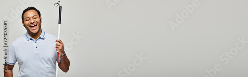 glad indian blind man in blue tee shirt holding walking stick and posing on gray backdrop, banner