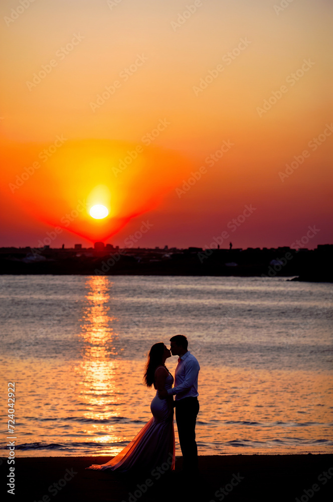 Fototapeta premium A man and pregnant woman kiss on a beach as the sun sets. ai generative