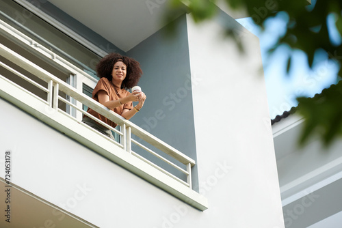 Happy Latin woman standing on balcony with cup of morning coffee