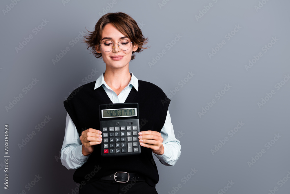 Portrait of satisfied girl with bob hairdo dressed waistcoat in eyewear look at calculator in arms isolated on gray color background Portrait of satisfied girl with bob hairdo dressed waistcoat in eyewear look at calculator in arms isolated on gray color background