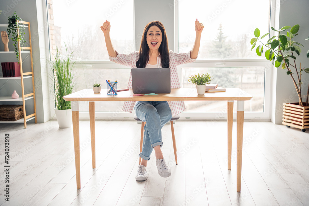 Full length photo of overjoyed cheerful person sit chair raise fists attainment desk laptop spacious office indoors Full length photo of overjoyed cheerful person sit chair raise fists attainment desk laptop spacious office indoors