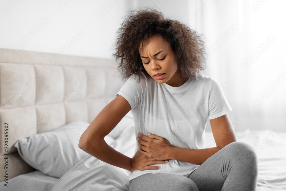Unhappy black young lady clutches her abdomen sitting on bed