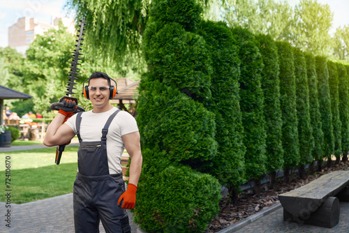 Muscular male gardener in uniform, safety glasses and gloves holding electric hedge trimmer in hands. Handsome caucasian man smiling and looking at camera while standing on back yard.