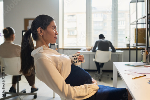 Young bored pregnant businesswoman in smart casualwear having tea or coffee while sitting in front of computer screen in office
