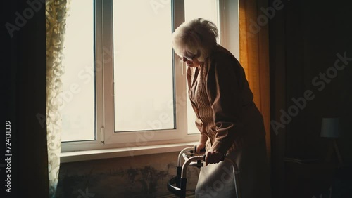 Sad elderly woman standing with a walking frame near the window, feeling lonely photo