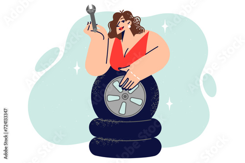 Woman auto mechanic leans over wheels and demonstrates wrench for self-repairing car