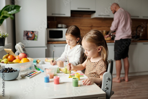 Adorable little girls sitting by kitchen table and painting with gouache and watercolors while their father preparing breakfast for family photo