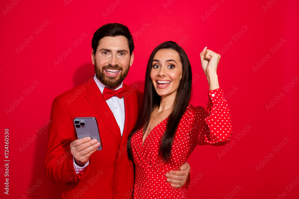 Photo of two attractive delighted lovers embrace hold smart phone raise fist empty space isolated on red color background