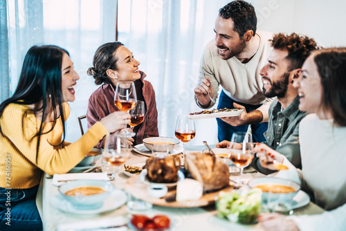Happy group of friends having dinner party at home - Cheerful young people having lunch break together - Life style concept with guys and girls celebrating thanksgiving - Man serves vegetables photo