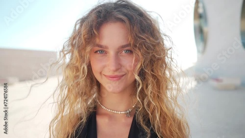 Close-up portrait of smiling blond woman looking directly at camera. Attractive female model with long hair slightly tousled by wind, portraying happy and confident expression on face.