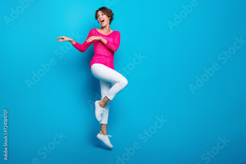 Full length photo of funky carefree woman wear knitted neon shirt jumping high dancing empty space isolated blue color background