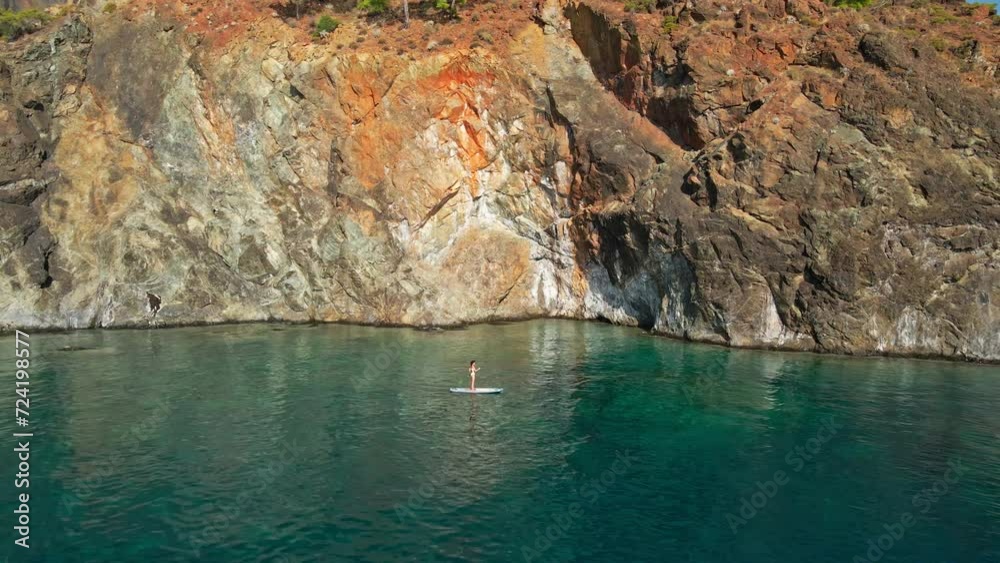 Aerial view of woman paddling a stand-up paddleboard or SUP board on a calm sea along rocky shore Aerial view of woman paddling a stand-up paddleboard or SUP board on a calm sea along rocky shore