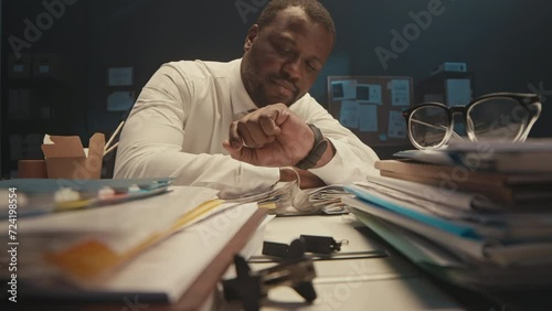 Stab shot of exhausted Black businessman sleeping at office desk with stacks of documents in folders on it, working overtime at night meeting deadlines photo