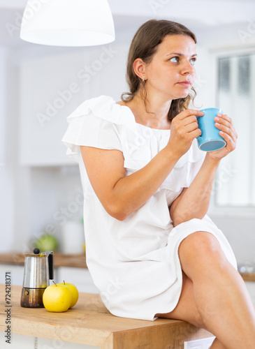Portrait of relaxed young woman sitting on kitchen countertop, drinking coffee or tea in morning