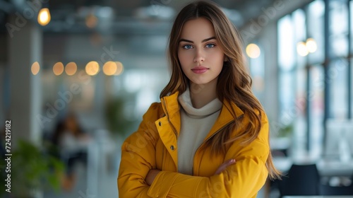 A young pretty woman in a yellow jacket stands in a modern office with her arms crossed