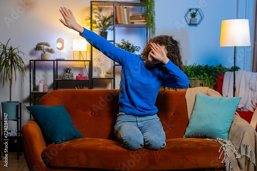 Excited young woman with curly hair having fun dancing and moving to rhythm sitting on sofa in living room. Cheerful female girl making dub dance gesture in apartment. Trendy girl dabbing at home. photo