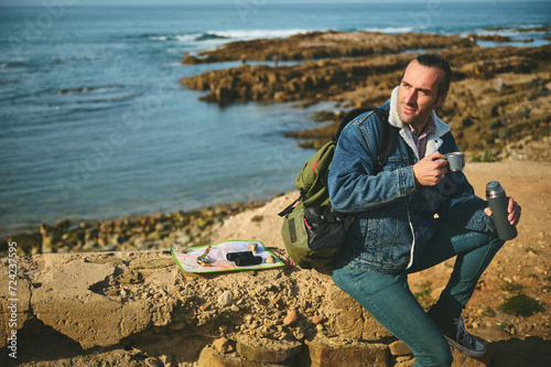 Slika na platnu Happy adventurer sitting by Atlantic beach, smiles looking aside, holds a thermo