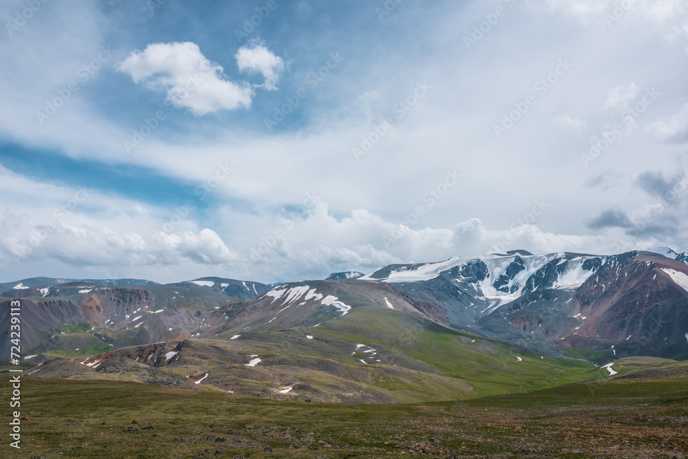 Fototapeta premium Dramatic alpine view from stony grassy hill to large snow-capped mountain range under cloudy gray sky. Beautiful mountain landscape with green rock hills against high mountains in changeable weather.