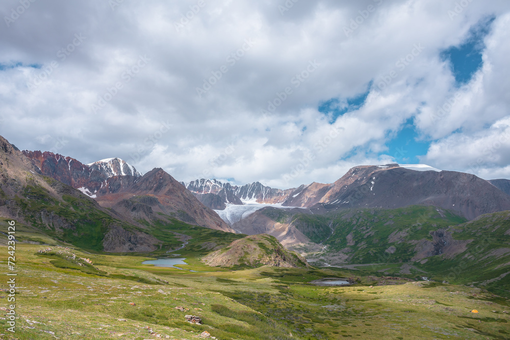 Colorful scenery with two beautiful lakes among green hills and rocks in alpine valley with view to big glacier and large snow mountain range far away under clouds in blue sky. High snowy mountains.